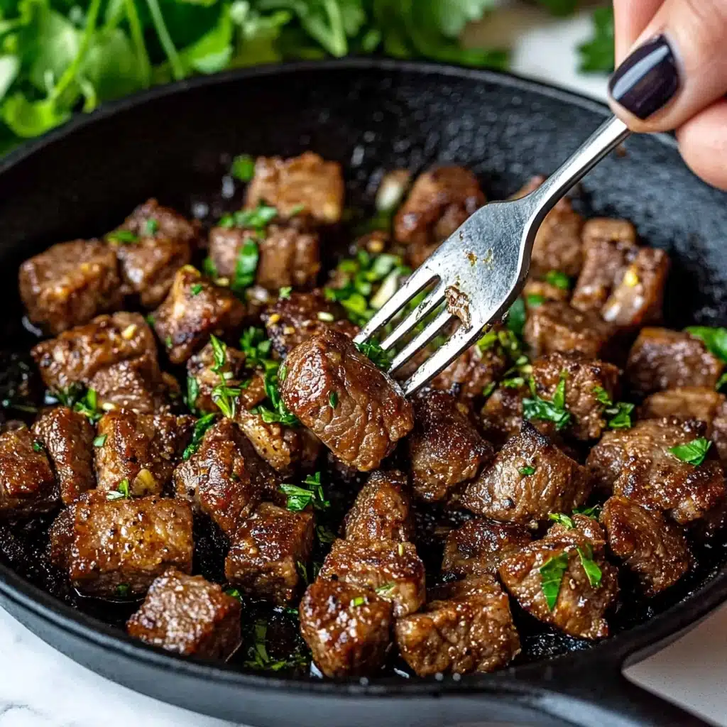 Garlic Butter Steak Bites, Steak Bites Recipe, Easy Steak Dinner, Quick Steak Appetizer, Tender Steak Bits A close-up of browned meat cubes in a black cast iron pan, each piece sprinkled with chopped green herbs and small bits of garlic, showing a mix of crispy and tender textures; a woman's hand with dark painted nails holds a silver fork piercing one meat cube, the background softly blurred with green leaves, and the pan sits on a white marbled surface.