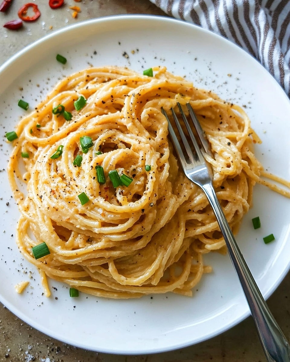 Creamy Cajun Linguine, Cajun pasta with creamy sauce, spicy Cajun linguine recipe, quick Cajun pasta dish, easy Cajun pasta meal A white plate holds a mound of spaghetti noodles coated in a creamy, light orange sauce, sprinkled with small green chive pieces and black pepper flakes. The spaghetti strands form two main clusters: a larger one on the left and a smaller, twirled nest on a fork placed on the right side of the plate. The fork's handle extends off the bottom edge of the plate. The plate rests on a brown surface with scattered red chili flakes and chopped onion bits around it. A striped cloth napkin peeks from the top right corner. The