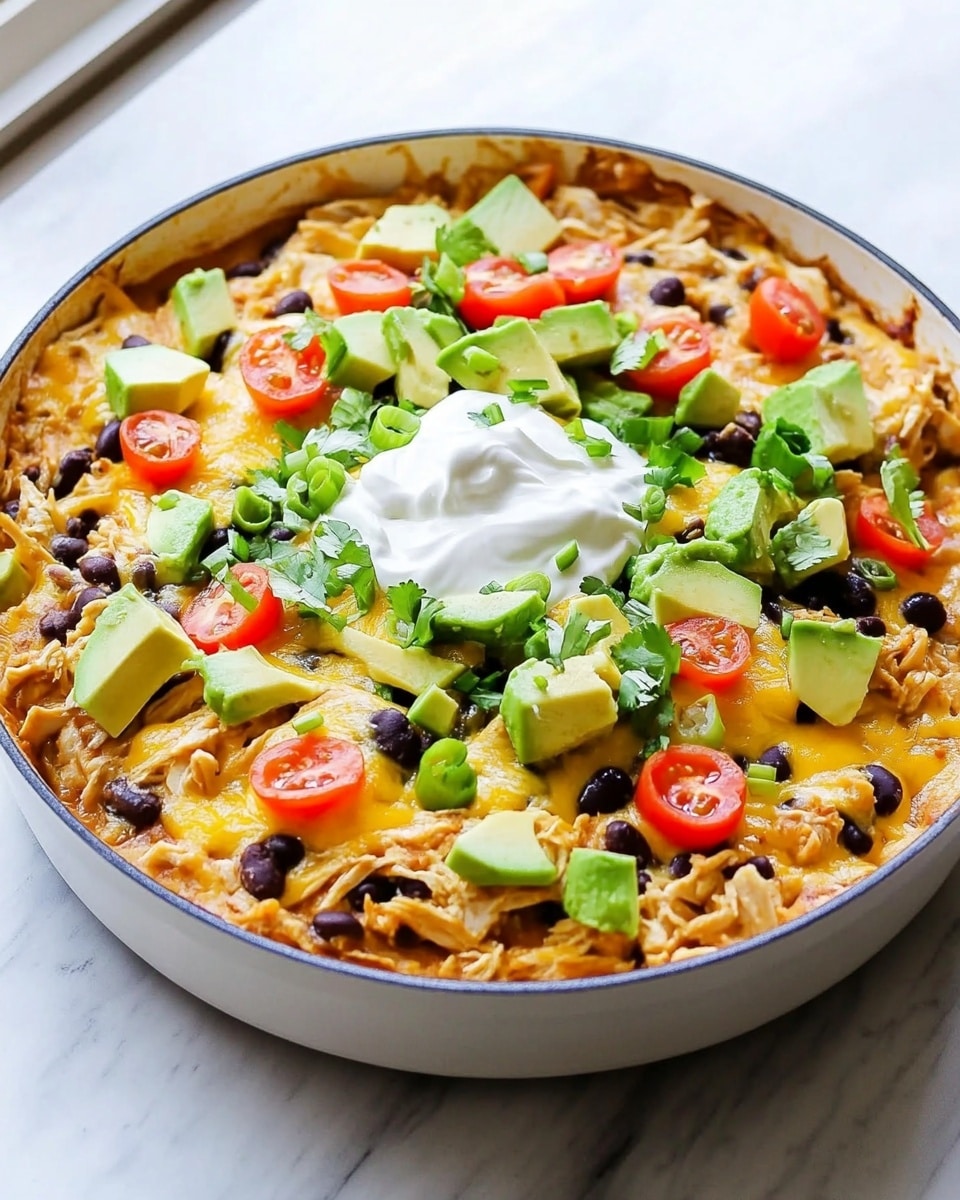 Chicken Black Bean Enchilada Skillet, quick chicken skillet dinner, easy Mexican chicken recipe, one-pan enchilada skillet, simple weeknight chicken dish The dish is a creamy, cheesy skillet meal served in a white pan with two handles, placed on a white marbled surface. The base layer is a warm, orange-colored sauce covering pasta or tortillas mixed with black beans, giving a textured look with bits peeking through. Melted white and yellow cheese spreads evenly over the top, slightly browned in some areas, creating a rich, gooey layer. Cherry tomato halves and small avocado cubes are scattered generously on top, adding bright red and green pops of color. There is a dollop of smooth white sour cream in the center, surrounded by finely chopped green onions sprinkled evenly over the entire dish. The light from a nearby window highlights the glossy cheese and fresh toppings, making the dish look fresh and hearty.