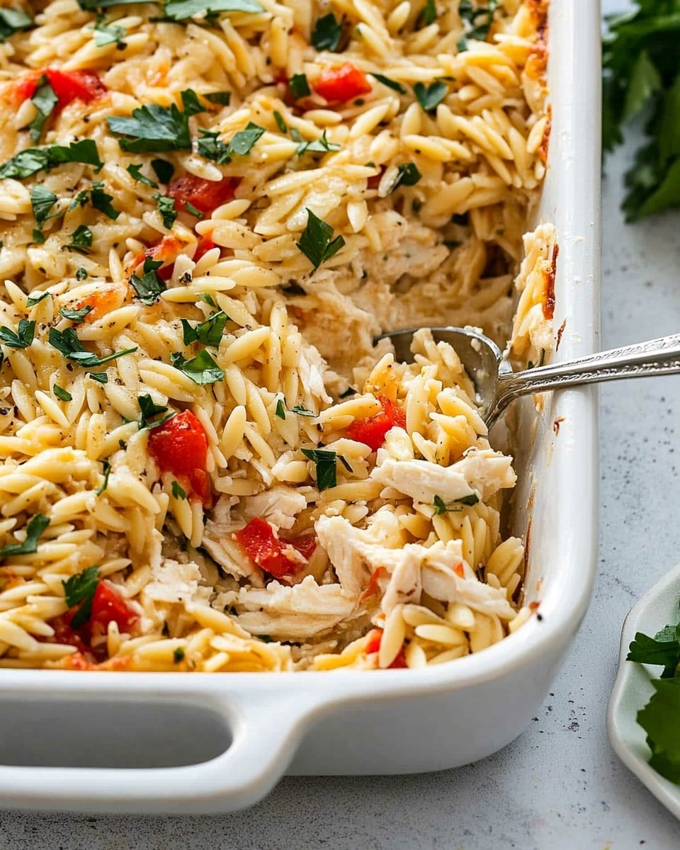 Greek Chicken and Orzo Casserole, Mediterranean chicken bake, easy Greek chicken casserole, healthy chicken and orzo dish, flavorful baked chicken casserole The image shows a close-up of a white rectangular baking dish filled with a mixed pasta casserole. The dish has one visible thick handle on the near side. The casserole is full of small pasta pieces mixed with shredded light-colored chicken, chopped red bell peppers, leafy green herbs, and small bits of white cheese sprinkled on top. The texture looks creamy with a mix of soft and slightly chunky ingredients. A silver spoon is partly inserted into the casserole on the right side. The dish is placed on a white marbled surface with a small white dish holding some green leaves partially visible on the right edge.