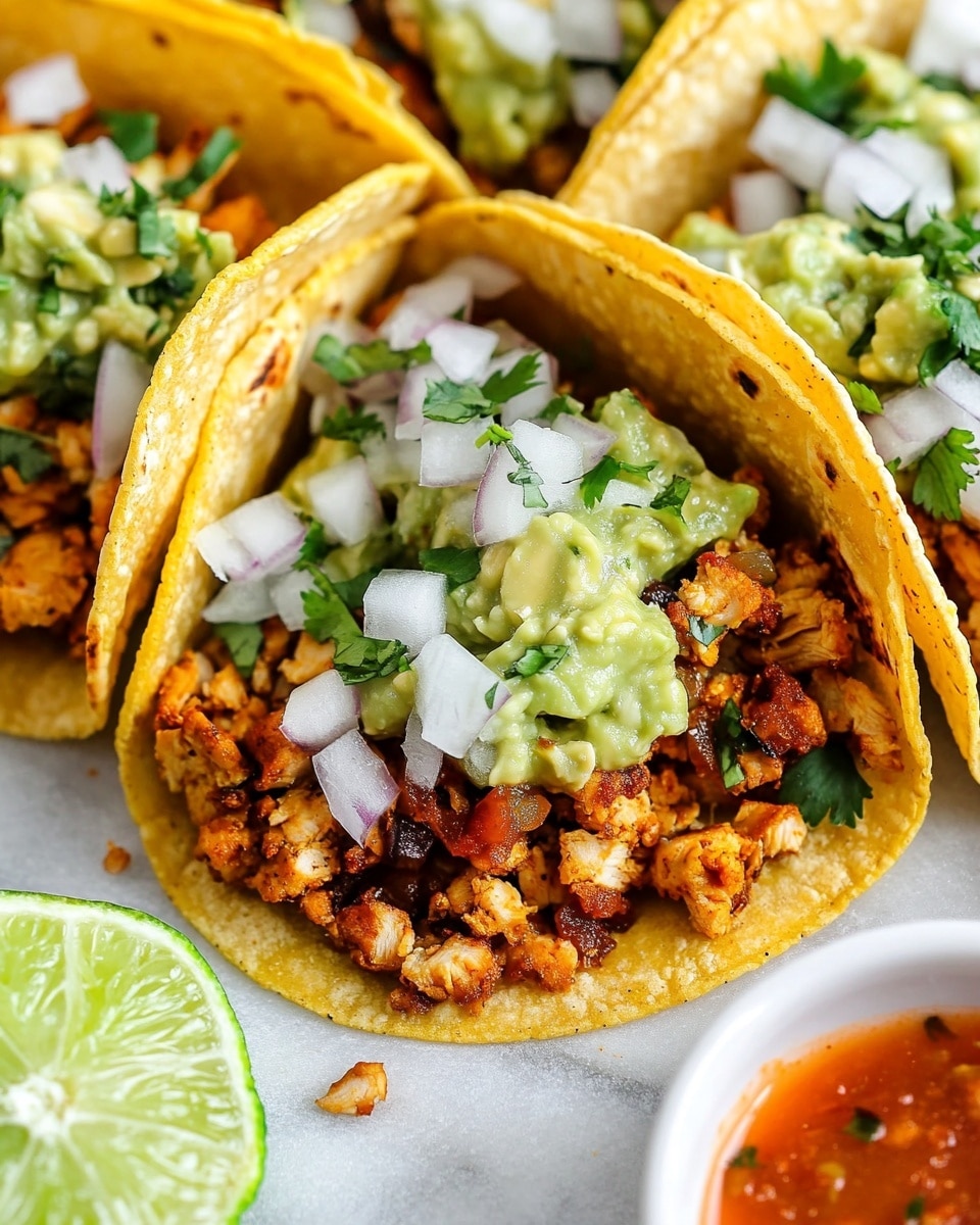 Chicken Street Tacos Onion Salsa, chicken street tacos, Mexican chicken tacos, easy street tacos recipe, flavorful taco recipes Three tacos are shown close together on a white marbled surface, each with a soft yellow corn tortilla as the base, lightly toasted with slight browning spots. Inside the tortillas are layers starting with small pieces of cooked, seasoned meat that is reddish-brown. On top of the meat, there is a mix of white chopped onions and green cilantro, giving a fresh and colorful look. A dollop of green guacamole sits on one side of each taco, adding a creamy texture. In the background, a small round white bowl holds a reddish sauce, slightly blurred to keep focus on the tacos. The photo was taken with an iphone