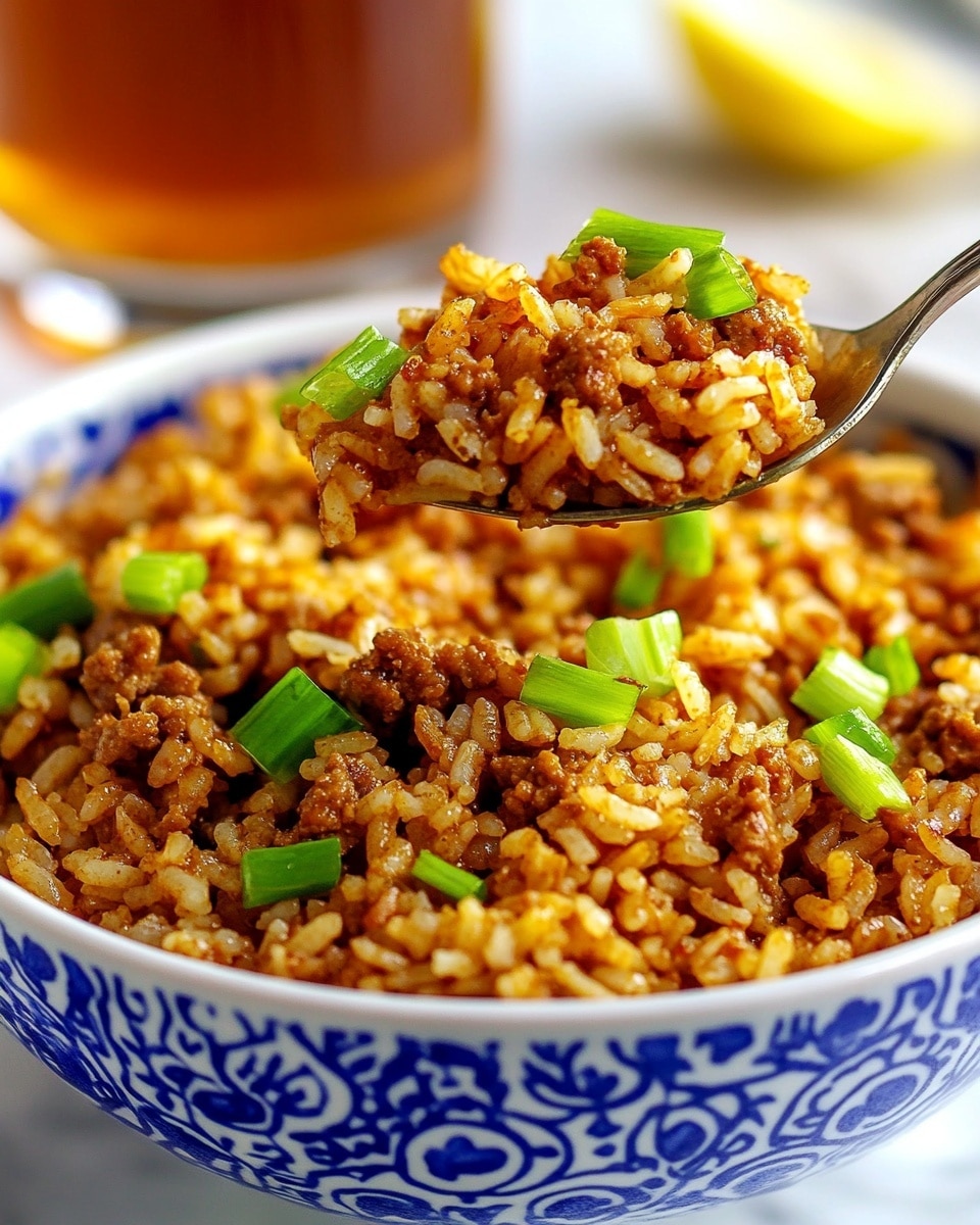 Easy Cajun Dirty Rice with Sausage, Cajun dirty rice, Cajun sausage rice, flavorful dirty rice recipe, quick Cajun rice dish A close-up of a white bowl with blue patterns filled with cooked rice mixed with browned ground meat and green chopped scallions scattered on top. A spoon lifted by a woman's hand holds some of the rice and meat mixture above the bowl. The rice looks fluffy with a reddish-brown color from spices, and the bowl is placed on a white marbled surface. In the background, there is a blurred glass of iced tea with a lemon wedge.