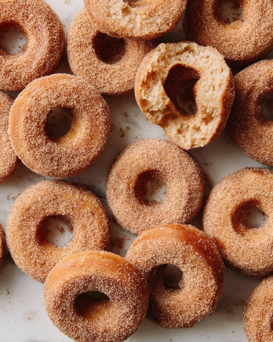 This image shows a close-up view of several small, round doughnuts with a hole in the center, all coated in cinnamon sugar. There are about ten doughnuts overlapping each other, with one doughnut in the middle broken into two pieces, showing a soft, light brown inside texture. The cinnamon sugar layer looks grainy and slightly uneven, covering each doughnut completely. The doughnuts are set on a white marbled surface, adding a clean background that highlights the warm brown and beige tones of the doughnuts. photo taken with an iphone --ar 4:5 --v 7