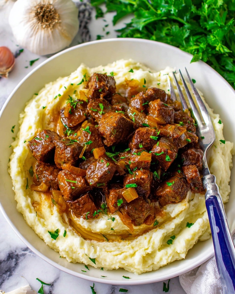 The image shows a white round plate filled with a base layer of creamy mashed potatoes that have a smooth and fluffy texture with slight swirls. On top, there is a generous layer of brown, glossy beef stew pieces, cut into small cubes and garnished with finely chopped green herbs. The beef looks tender and well-cooked, and there are small bits of caramelized onion mixed in. To the side, a fork with a blue handle rests on the plate, and in the background, there are green leafy herbs and a whole garlic bulb on a white marbled surface. photo taken with an iphone --ar 4:5 --v 7