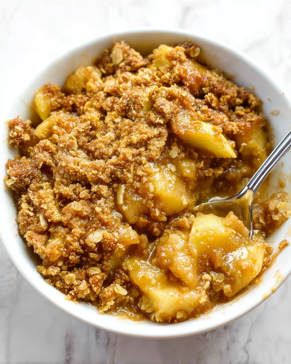 The dish is a white bowl filled with a warm apple crumble dessert. There are three visible layers: the bottom layer is a soft, gooey light brown apple filling with chunks of apple and a syrupy texture; above it is a thick, golden brown crumb topping made of oats and sugar that looks crispy and crunchy; a silver spoon is placed inside the bowl slightly to the right. The background is a white marbled surface. photo taken with an iphone --ar 4:5 --v 7