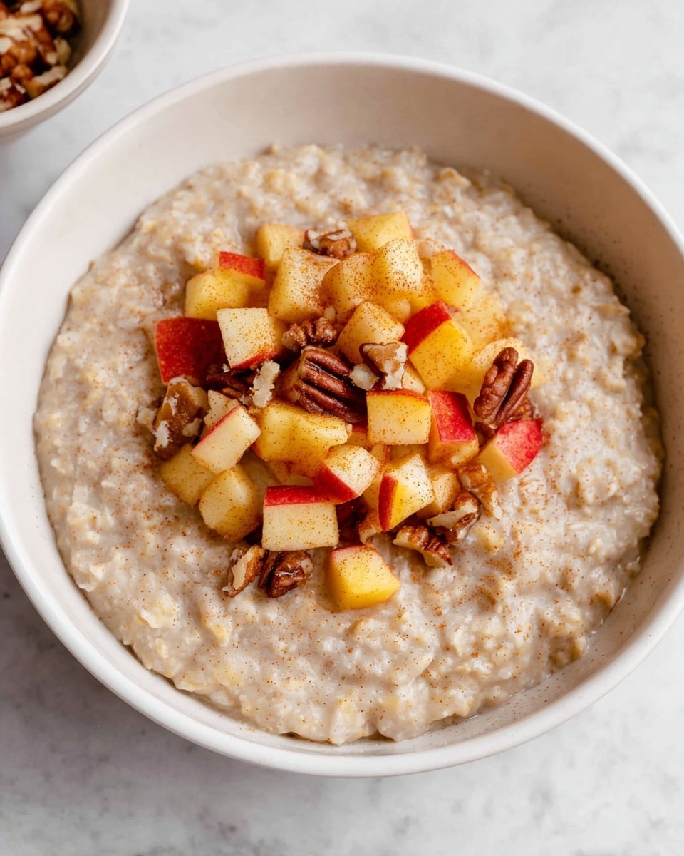 A white bowl filled with one layer of creamy oatmeal that looks soft and thick, showing a pale beige color and grainy texture. On top at the center, a second layer of small diced apple pieces in red and yellow colors mixed with brown pecans, giving a contrast with their shiny and rough textures. The bowl is set on a white marbled surface. The photo taken with an iphone --ar 4:5 --v 7