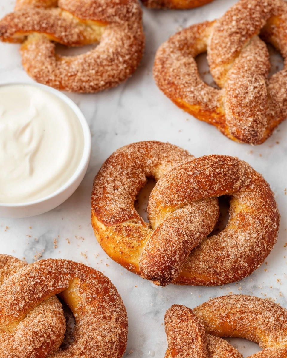 The image shows several soft pretzels covered evenly with a brown cinnamon sugar coating, arranged on a white marbled surface. Each pretzel has a twisted knot shape with a slightly golden baked texture beneath the cinnamon sugar layer. In the bottom left corner, there is a white bowl filled with smooth, creamy white dipping sauce. The background is bright and clean, enhancing the warm color of the pretzels. photo taken with an iphone --ar 4:5 --v 7