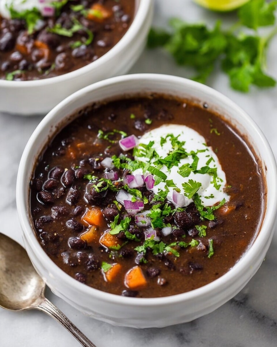 A white bowl filled with thick black bean soup showing layers of whole black beans and small pieces of orange carrot in a dark, smooth broth. On top, there is a dollop of white sour cream, sprinkled with chopped fresh green cilantro and small pieces of purple onion. The bowl sits on a white marbled surface next to a vintage silver spoon and fresh green cilantro leaves. Another white bowl with the same soup is partially visible in the background. Photo taken with an iphone --ar 4:5 --v 7