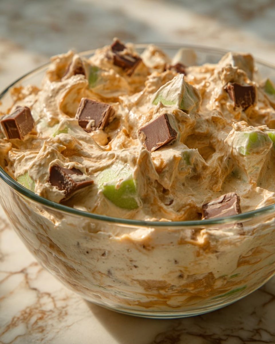 A clear glass bowl filled with a creamy, light brown mixture that has a whipped texture and visible chunks of green apple and pieces of a chocolate candy bar mixed throughout. The bowl sits on a white marbled surface, and the colors are warm and inviting with soft lighting highlighting the smooth and chunky elements of the dish. The mixture looks fluffy with the chunks scattered evenly on top and within. Photo taken with an iphone --ar 4:5 --v 7