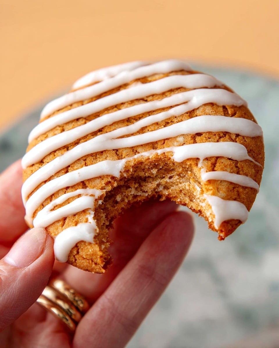 A close-up of a round cookie with a golden brown top covered in evenly spaced white icing lines. The cookie has a visible soft inside that is lighter in color. A woman's hand with a gold ring on the ring finger is holding the cookie, which has a bite taken out of it. The background shows a white marbled surface under soft warm light. photo taken with an iphone --ar 4:5 --v 7