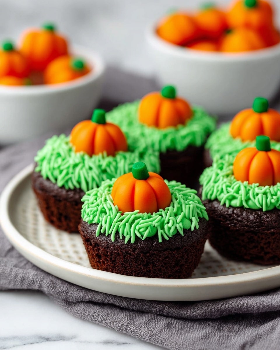 Four small brown cupcakes are on a round white plate with a ridged edge. Each cupcake has a top layer of bright green frosting piped to look like grass. On top of the green frosting, there is an orange pumpkin-shaped candy with a small green stem. In the background, there are two white bowls, one filled with more pumpkin candies and the other with candy corn. The plate is placed on a gray cloth on a white marbled surface. Photo taken with an iphone --ar 4:5 --v 7