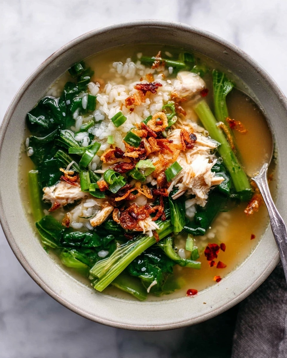 A bowl of soup with clear broth fills a light gray ceramic bowl, placed on a white marbled surface. Inside the bowl, there are soft white rice grains mixed with tender white chicken pieces and bright green leafy vegetables with stalks. On top, small crispy golden-brown fried shallots are scattered along with some drops of red chili oil adding color contrast. A silver spoon is partly submerged on the right side of the bowl. Photo taken with an iphone --ar 4:5 --v 7