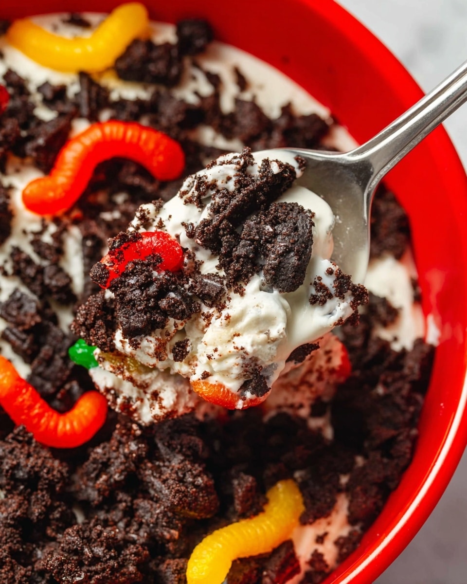 A close-up image shows a red bowl filled with dark brown crushed cookie crumbs as the base layer, topped with colorful gummy worms in orange, yellow, and red scattered around. A silver spoon is scooping up a portion of white creamy layer covered with chunks of dark cookie crumbs, mixing the creamy texture with crunchy bits. The background is a white marbled texture, and the focus is on the spoonful of the layered dirt dessert with cookie pieces and gummy worms visible. photo taken with an iphone --ar 4:5 --v 7