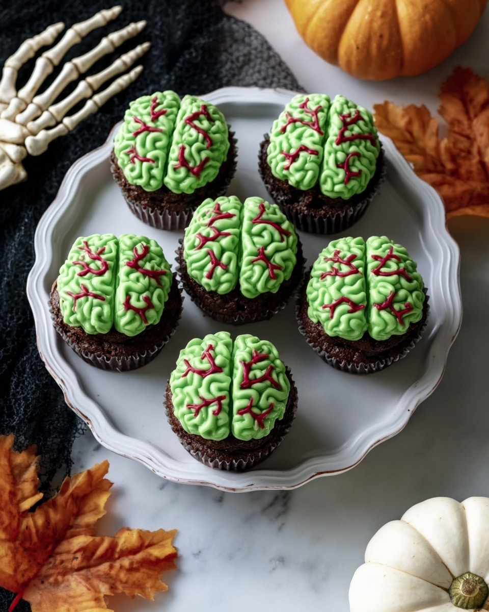 Five cupcakes are placed in a circle on a white plate with a wavy edge. Each cupcake has two layers: a chocolate base and a green frosting top shaped like a brain, with thin red lines running through the green frosting to show veins. The plate is on a white marbled surface, with a skeleton prop’s woman’s hand resting near the plate on the left side, a white pumpkin in the bottom right, and a few orange autumn leaves scattered around. Photo taken with an iphone --ar 4:5 --v 7