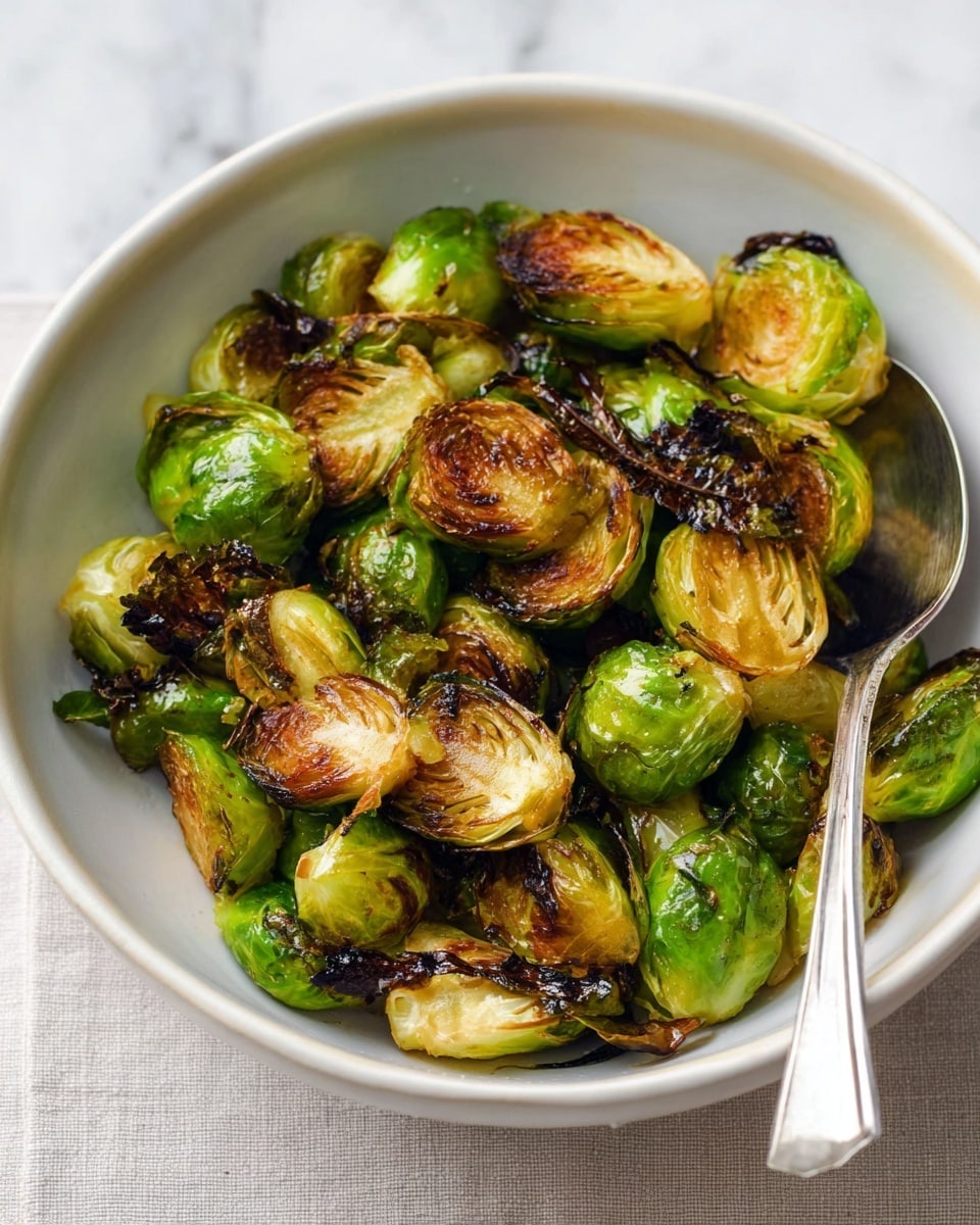 A white bowl fills the image, holding roasted Brussels sprouts that are halved, showing a mix of bright green and golden brown layers with some crispy, darkened edges for texture. The Brussels sprouts have a slightly shiny surface from being cooked in oil, with varied shades of green and brown giving a roasted look. Inside the bowl, a silver metal spoon rests partially in the Brussels sprouts on the right side. The bowl is placed on a white marbled texture that contrasts softly with the green and brown colors of the vegetables. photo taken with an iphone --ar 4:5 --v 7