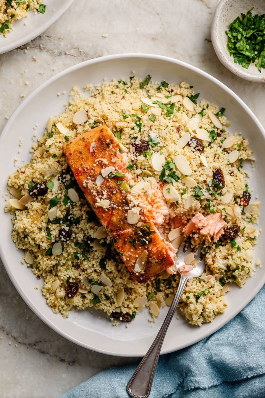 A white bowl filled with a base layer of light yellow couscous mixed with chopped green herbs and small dark raisins. On top of the couscous, there is a piece of cooked salmon with a golden brown crust, broken open to show the soft pink inside. Thin almond slices and extra chopped green herbs are scattered on the dish. A silver spoon rests on the right side of the bowl, partially under the couscous. The bowl sits on a white marbled surface, and a light blue cloth napkin is visible nearby. photo taken with an iphone --ar 2:3 --v 7 - Moroccan-Spiced Salmon with Citrus Couscous, Moroccan salmon recipe, quick salmon dinner, citrus couscous recipe, flavorful weeknight salmon