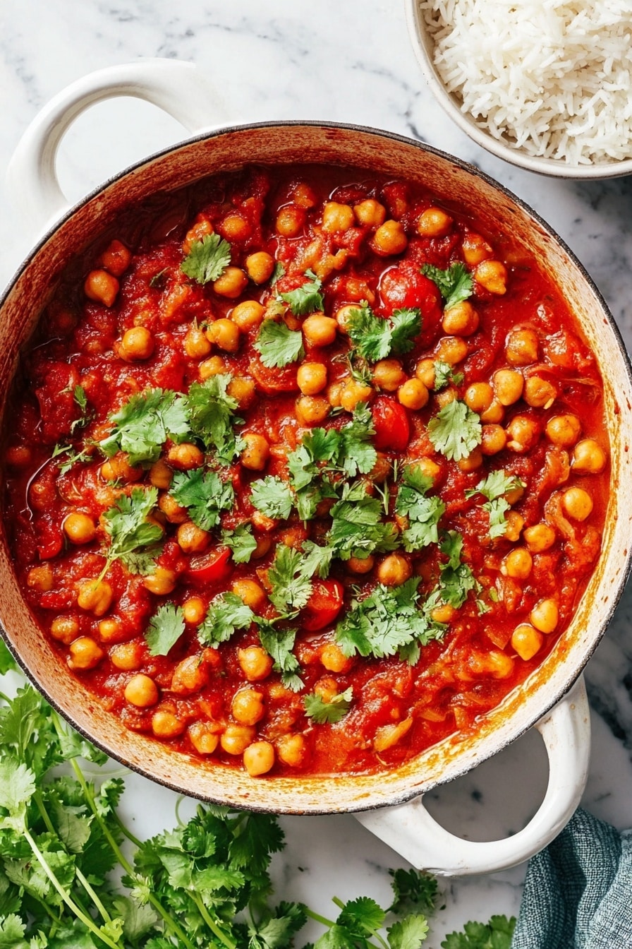 A white cast iron pan filled with a one-layer chickpea stew that has a rich red tomato sauce mixed with visible tomato chunks. The stew is topped with scattered fresh green cilantro leaves. The pan rests on a white marbled surface with some loose cilantro at the bottom left. A small white bowl with cooked white rice is visible on the top right corner. The photo taken with an iphone --ar 2:3 --v 7 - Easy Chana Masala, vegan chickpea curry, spicy Indian chickpea dish, quick vegan curry, wholesome chickpea recipe