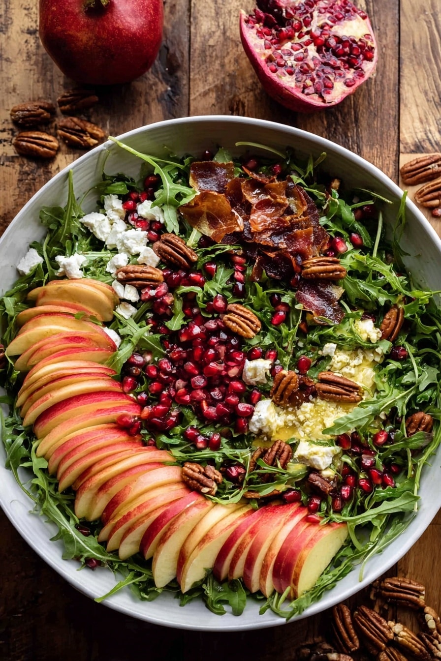 A large white bowl holds a colorful salad layered with fresh green arugula leaves spread across the bottom and middle. Thin slices of red and yellow apple are arranged in slightly fanned rows all around the bowl on top of the greens. Bright red pomegranate seeds are scattered generously over the salad, adding vibrant contrast. Small white crumbles of cheese sit in patches on one side of the bowl. Toasted pecans and nuts, dark brown in color, are mixed through the salad, adding texture. There are thin, crispy brown slices of prosciutto or a similar cured meat placed lightly on the salad. The bowl sits on a rustic wooden surface with halved pomegranate and small pecans nearby. Photo taken with an iphone --ar 2:3 --v 7 - Autumn Harvest Honeycrisp Apple and Feta Salad, fall apple salad with feta and nuts, Easy autumn fruit salads, seasonal apple and cheese salad, healthy fall salad recipes
