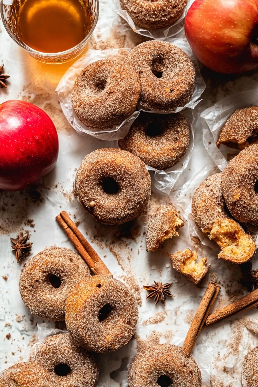 The image shows many cinnamon sugar donuts placed closely on a white marbled surface. Most donuts are whole, with a rough texture covered in sugar and cinnamon. A few donuts are broken, revealing a soft, golden inside with a slightly crumbly texture. Some donuts are wrapped in thin, translucent white paper, adding layers and depth. Near the donuts, there is a bright red apple and a clear glass cup filled with light brown tea and cinnamon sticks inside. Star anise is scattered around for decoration. The scene looks warm and cozy with soft natural light. Photo taken with an iphone --ar 2:3 --v 7 - Baked Cinnamon Crunch Apple Cider Doughnuts, baked apple cider doughnuts, cinnamon sugar doughnuts, fall baked doughnuts, easy oven baked doughnuts