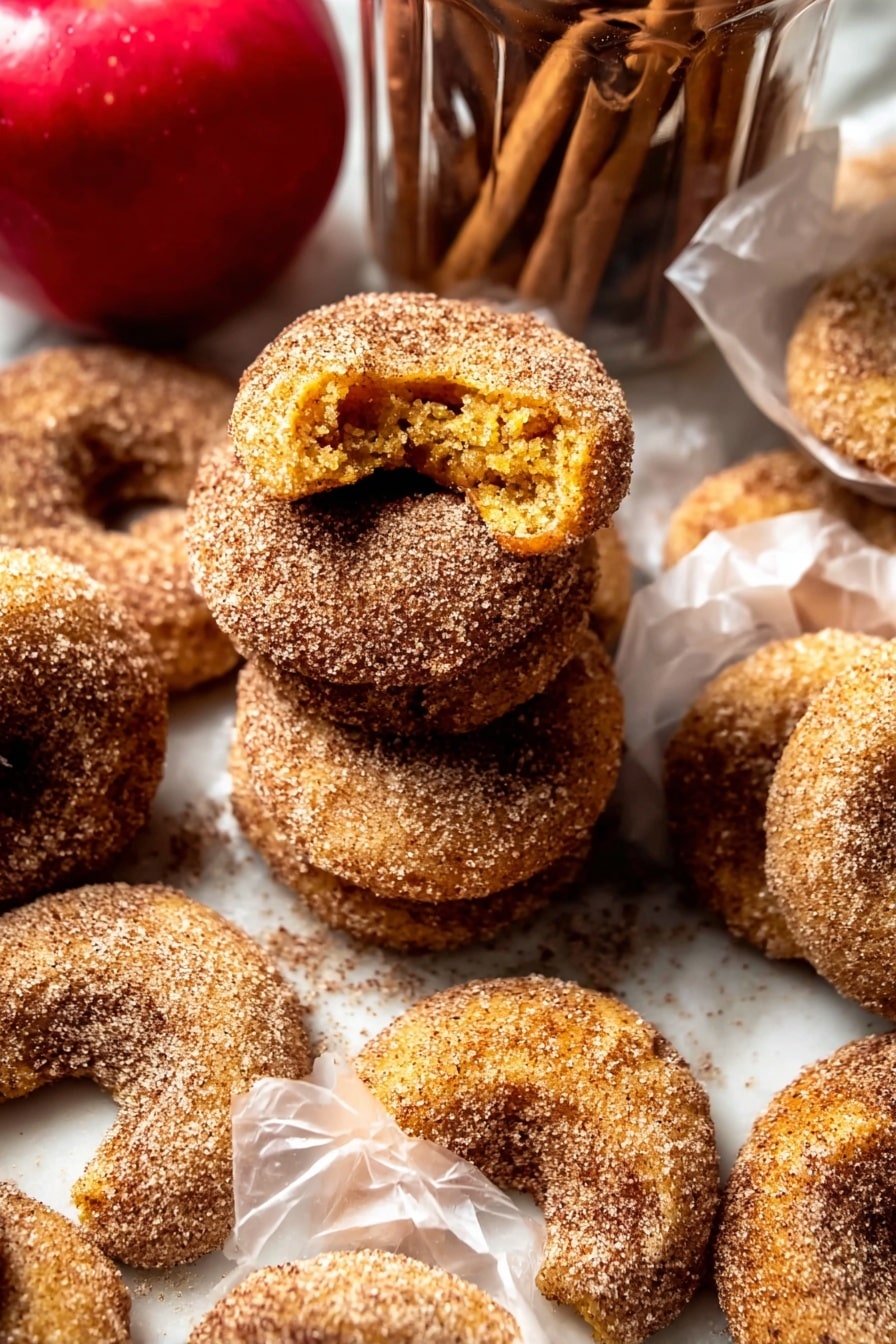 The image shows several crescent-shaped cookies covered in a coarse layer of cinnamon sugar. The cookies are golden brown with a slightly rough texture beneath the sugar coating. Some cookies are stacked in a loose pile, while others are individually wrapped in translucent white paper. The cookies have a crumbly look, with one cookie broken open to reveal a soft yellow interior. In the background, there is a bright red apple and a clear glass jar filled with more cookies and cinnamon sticks on top. The setting uses a white marbled surface underneath all the items. photo taken with an iphone --ar 2:3 --v 7 - Baked Cinnamon Crunch Apple Cider Doughnuts, baked apple cider doughnuts, cinnamon sugar doughnuts, fall baked doughnuts, easy oven baked doughnuts