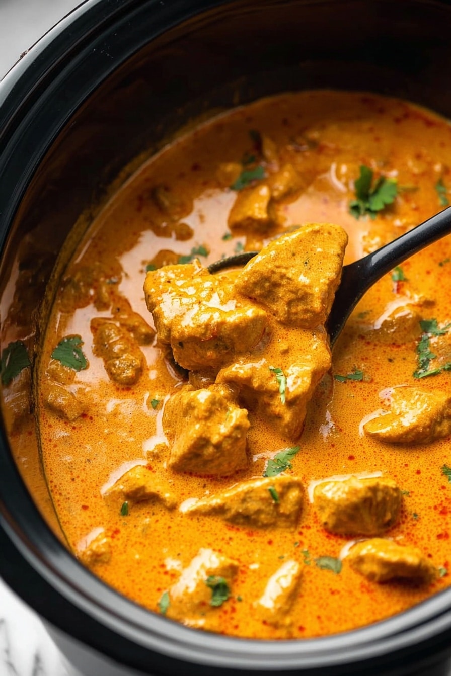This image shows a close-up of a creamy orange curry with chunks of tender meat in a black slow cooker. The curry sauce is thick and smooth with a slightly shiny surface. Small green herb pieces are sprinkled on top, adding a fresh contrast to the warm orange sauce. A black spoon is partially submerged in the curry on the right side, lifting some pieces of the meat. The background is a white marbled texture. photo taken with an iphone --ar 2:3 --v 7 - Slow Cooker Butter Chicken, Indian Butter Chicken, Easy Slow Cooker Dinner, Creamy Chicken Curry, Make-Ahead Indian Chicken
