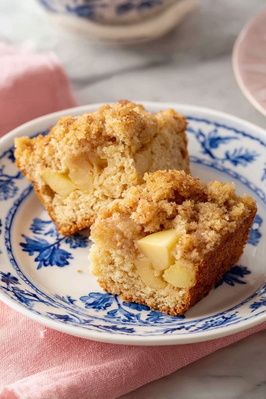 Two pieces of crumb-topped cake are placed on a white plate with blue floral patterns. The cake has a light brown color with soft, crumbly texture and visible chunks of yellow apple inside. The crumb topping is thicker and golden brown. The plate sits on a pink cloth, all set on a white marbled surface. Photo taken with an iphone --ar 2:3 --v 7 - Apple Cinnamon Muffins, Apple Cinnamon Muffins Recipe, Easy Apple Muffins, Fall Muffins, Moist Cinnamon Muffins