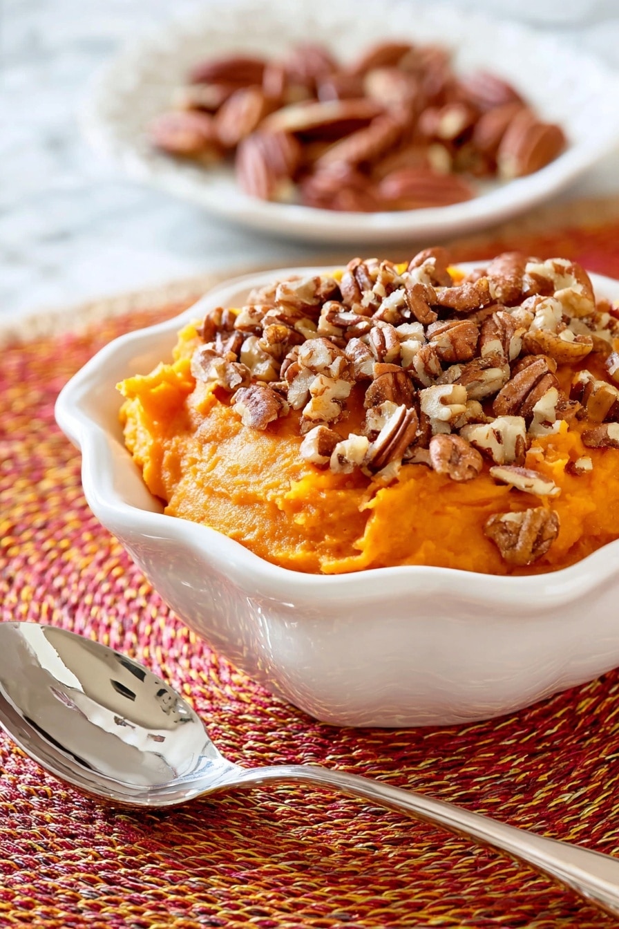 This image shows a white bowl filled with a thick, mashed orange sweet potato layer that is roughly shaped with a soft texture. On top of this layer, there is a generous amount of chopped pecans scattered unevenly, adding a crunchy contrast with their brown, nutty pieces. The bowl sits on a woven orange, red, and yellow circular placemat, and in the blurred background, part of another white dish with light brown pecans can be seen resting on a white marbled surface. photo taken with an iphone --ar 2:3 --v 7 - Slow Cooker Sweet Potato Casserole with Maple Syrup and Toasted Pecans, sweet potato casserole, holiday side dish, easy slow cooker sweet potato recipe, cozy fall casserole