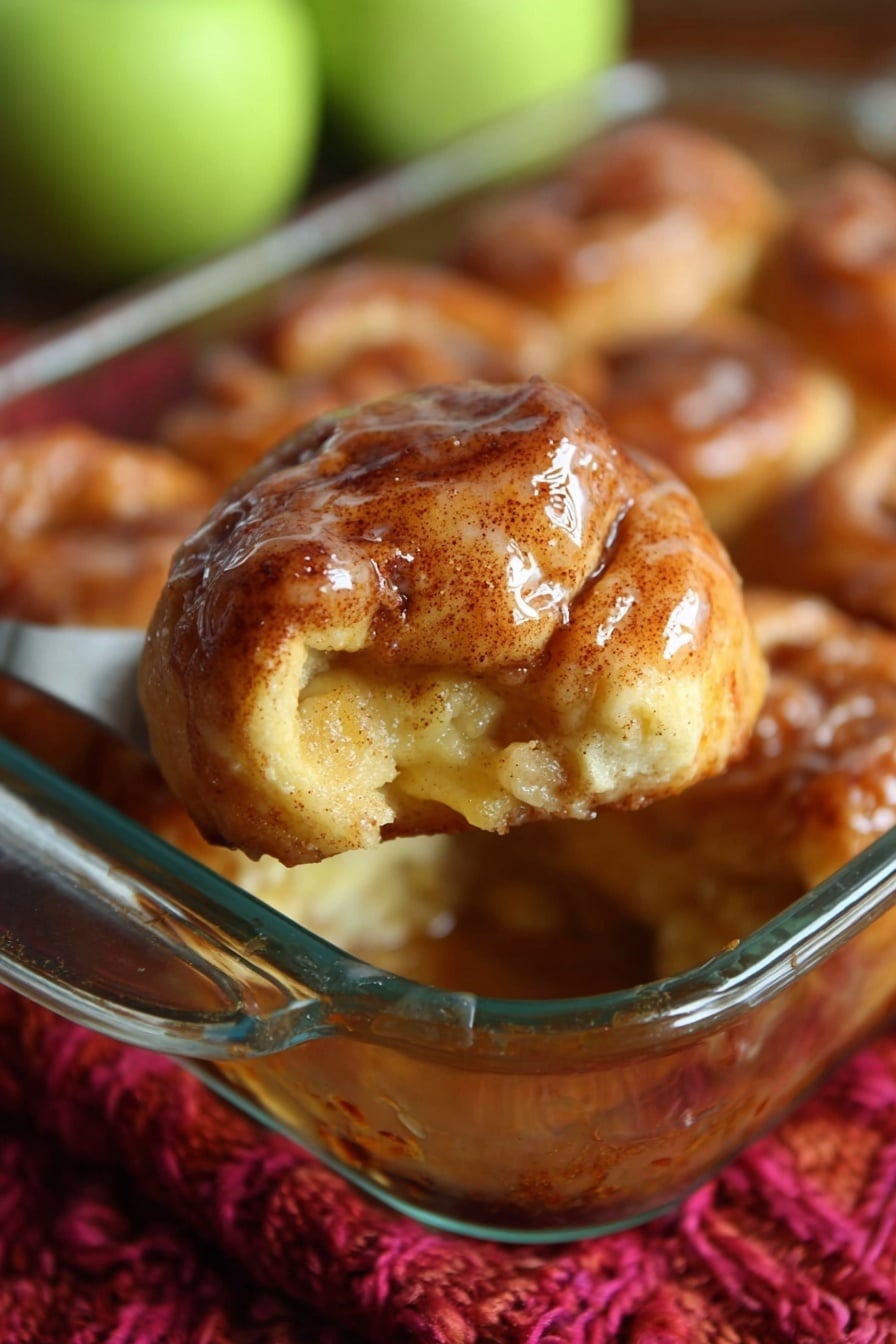 This image shows a close-up of a golden brown pastry with a shiny glaze, held above a glass baking dish filled with similar pastries. The pastry has a baked, slightly rough surface with visible cinnamon specks and a soft, filling visible at one side. The glass dish is resting on a thick, red knitted cloth, with blurred green apples in the background. photo taken with an iphone --ar 2:3 --v 7 - Apple Dumplings with Cinnamon and Sprite, apple cinnamon dessert, easy baked apple dumplings, fall dessert recipes, cinnamon apple treats