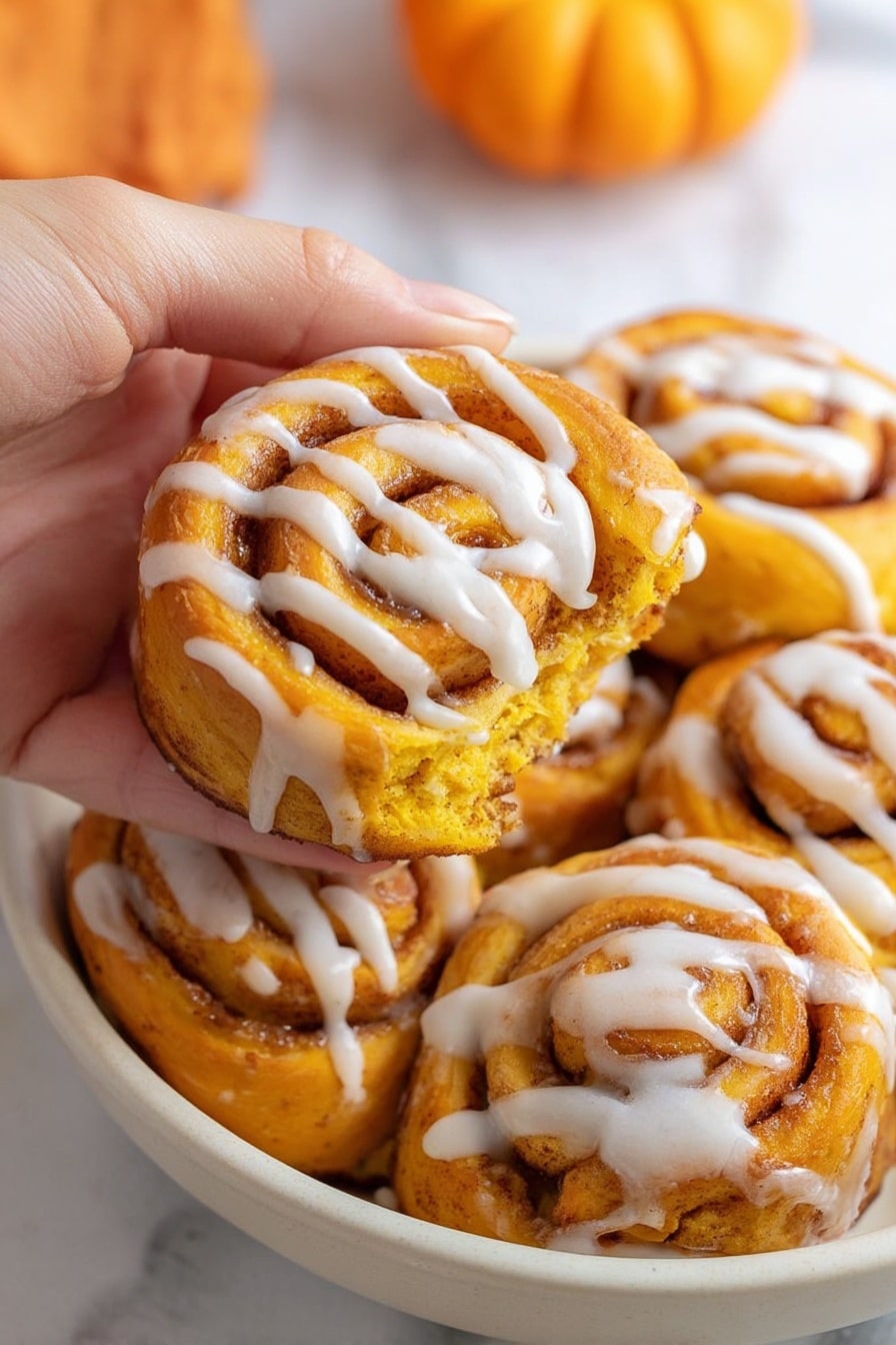 A woman's hand is holding one bright orange swirl roll with white icing drizzled across the surface, showing its soft and fluffy texture with cinnamon filling layers. Around it are several more identical rolls placed close together in a white bowl, all with the same orange color and white icing in spiral patterns. The background shows a white marbled surface with part of a small orange pumpkin visible on the right side. The overall scene highlights the warm, rich colors and creamy icing on the rolls. photo taken with an iphone --ar 2:3 --v 7 - Quick Pumpkin Cinnamon Rolls, easy pumpkin cinnamon rolls, cozy fall cinnamon roll recipe, quick autumn breakfast, pumpkin spice rolls