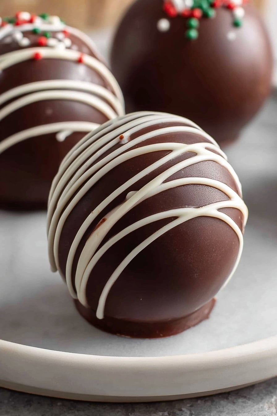 The image shows a smooth, shiny, dark brown chocolate sphere placed on a white plate with a white marbled texture under it. The sphere has thin white chocolate lines drizzled across its surface in a loose crisscross pattern, creating contrast and delicate texture on the curved chocolate shell. In the background, two more similar chocolate spheres are partially visible, each decorated with the same white drizzle and small dots of colored sprinkles. The overall look is clean and glossy, with the sphere’s curved layers clearly defined. photo taken with an iphone --ar 2:3 --v 7 - hot chocolate bombs, how to make hot chocolate bombs, homemade hot chocolate bombs, easy hot chocolate bombs recipe, chocolate sphere hot chocolate