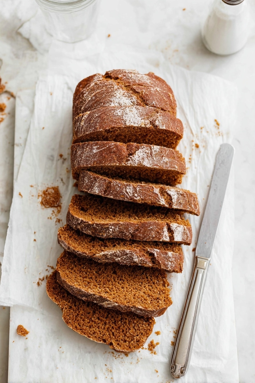 The image shows a loaf of brown bread cut into six thick slices, placed slightly overlapping each other on a white parchment paper. The bread has a textured crust with a few light flour spots on top and a soft, crumbly inside with a warm brown color. To the right of the bread, there is a silver knife with a simple handle resting on the parchment paper. The background is a white marbled surface with some bread crumbs scattered around, and the top right part of a clear glass and a white salt shaker are visible. Photo taken with an iphone --ar 2:3 --v 7 - Moist Pumpkin Bread, pumpkin bread recipe, fall pumpkin bread, easy pumpkin bread, tender pumpkin loaf