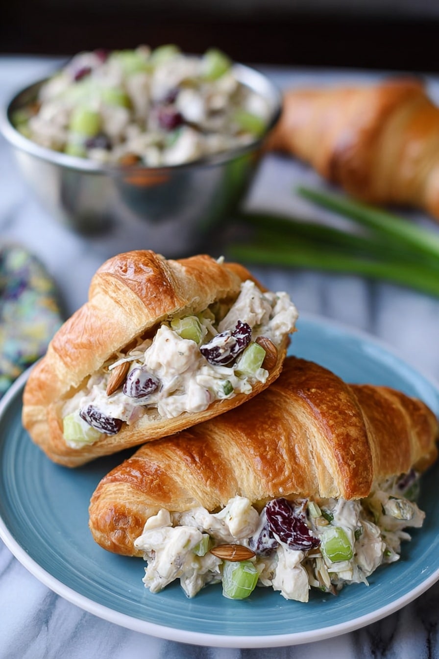 The image shows two croissants on a blue plate placed on a white marbled surface. One croissant is whole with a golden-brown, flaky texture. The other croissant is open and filled with a creamy salad made of small white chicken pieces, chopped celery, dried dark cranberries, sunflower seeds, and light beige dressing, layered inside the croissant’s light and flaky interior. In the background, there is a metal bowl filled with more of the salad, and some green onions and an additional croissant are blurred. Photo taken with an iphone --ar 2:3 --v 7 - Cranberry Chicken Salad, Cranberry Chicken Salad Recipe, Chicken Salad with Cranberries, Easy Cranberry Chicken Salad, Healthy Chicken Salad