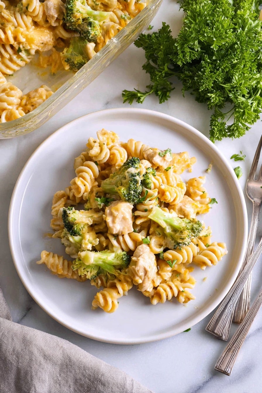 A white plate holds a serving of pasta made with spiral-shaped noodles mixed with chunks of light-colored cooked chicken and small, bright green broccoli florets, all coated in a creamy light yellow cheese sauce with a slightly shiny texture; the dish is placed on a white marbled surface next to a partially visible glass baking dish filled with more pasta, a small bunch of fresh parsley with deep green leaves, and a folded light gray cloth napkin holding three metal forks. Photo taken with an iphone --ar 2:3 --v 7 - Easy Chicken Noodle Casserole, chicken noodle casserole, creamy chicken pasta bake, family-friendly dinner recipes, simple casserole ideas