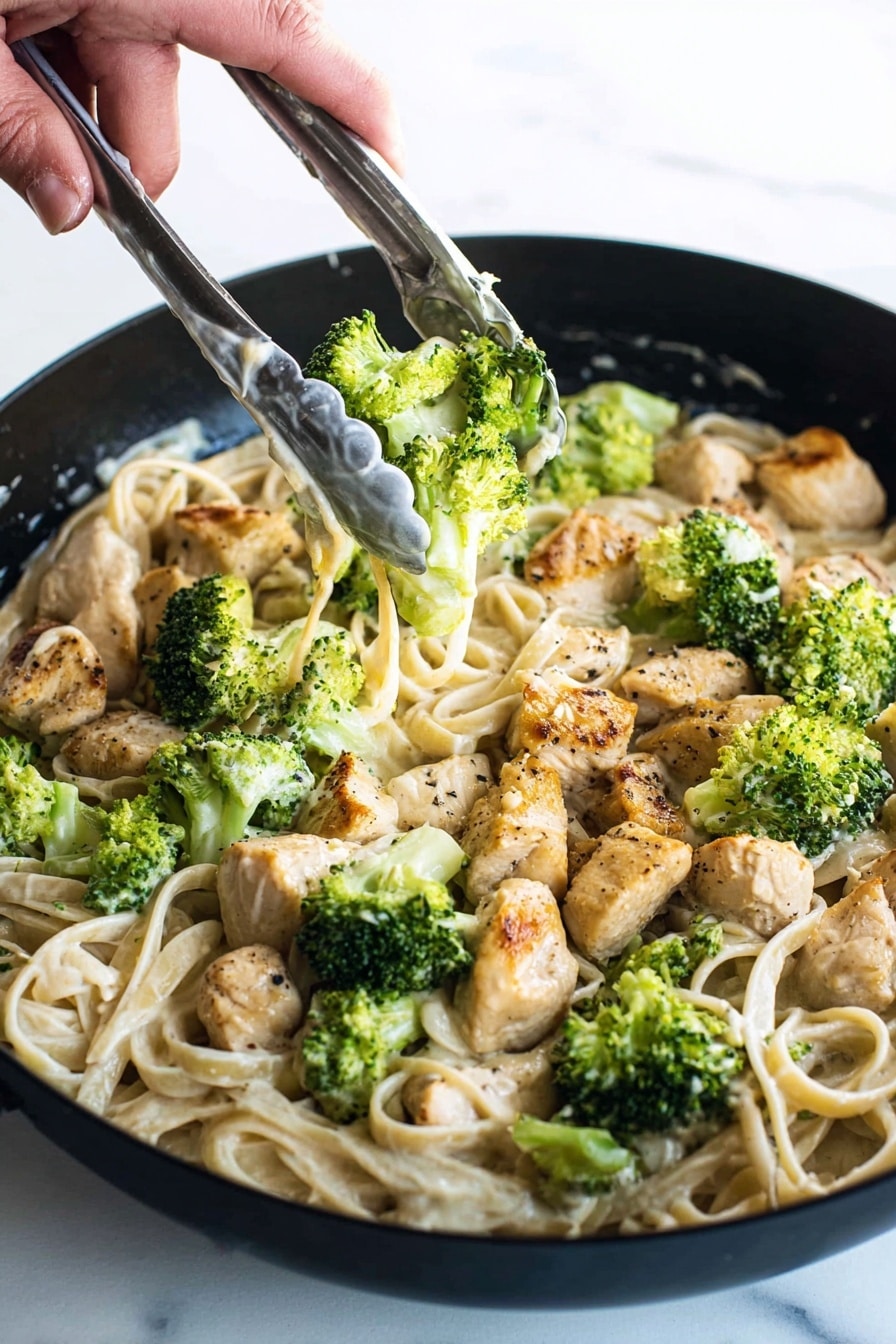 A close-up view of a black pan filled with creamy pasta layered at the bottom in a pale beige color with smooth texture. On top, there are chunks of cooked chicken pieces, golden brown with light black pepper spots, scattered evenly. Bright green broccoli florets, fresh and slightly steamed, are spread over the chicken and pasta. A woman's hand holding silver tongs with white sauce spots is seen in the upper right, picking up some broccoli and chicken. The background is a white marbled texture. photo taken with an iphone --ar 2:3 --v 7 - Quick Chicken Broccoli Fettuccine Alfredo, chicken broccoli pasta, creamy pasta dinner, easy weeknight chicken pasta, homemade Alfredo sauce