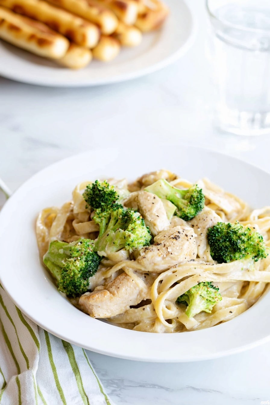 A white plate holds a serving of creamy fettuccine pasta mixed with light beige chicken pieces and bright green broccoli florets, all coated in a smooth, pale sauce with a few black pepper specks on top. In the background, out of focus, there is a white plate with golden brown breadsticks and a glass of water next to it. The scene is set on a white marbled surface with a folded white and green striped cloth visible beside the plate. photo taken with an iphone --ar 2:3 --v 7 - Quick Chicken Broccoli Fettuccine Alfredo, chicken broccoli pasta, creamy pasta dinner, easy weeknight chicken pasta, homemade Alfredo sauce