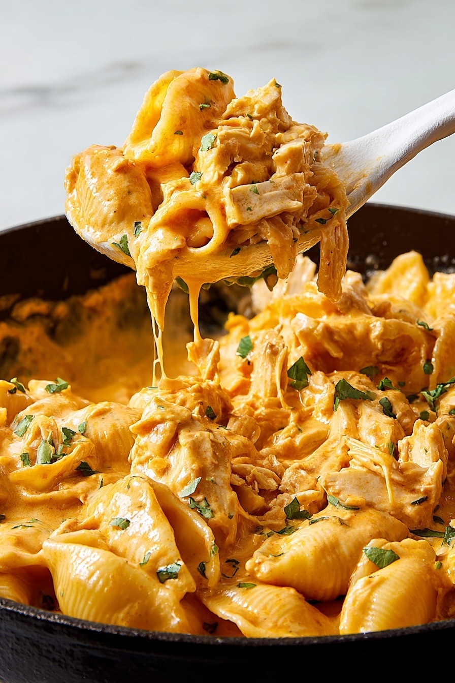 A close-up view of a black pan filled with creamy pasta shells covered in an orange cheese sauce. The dish shows layers of tender pasta shells mixed with shredded chicken, all coated thickly by the rich sauce. Small green herbs are scattered on top, adding spots of color. A white spoon lifts a portion of the creamy pasta and chicken from the pan, with the sauce dripping off the edge. The background is a white marbled texture. photo taken with an iphone --ar 2:3 --v 7 - White Chicken Chili Mac, White Chicken Chili Mac Skillet, creamy chicken skillet recipe, easy weeknight dinner ideas, cheesy chicken pasta