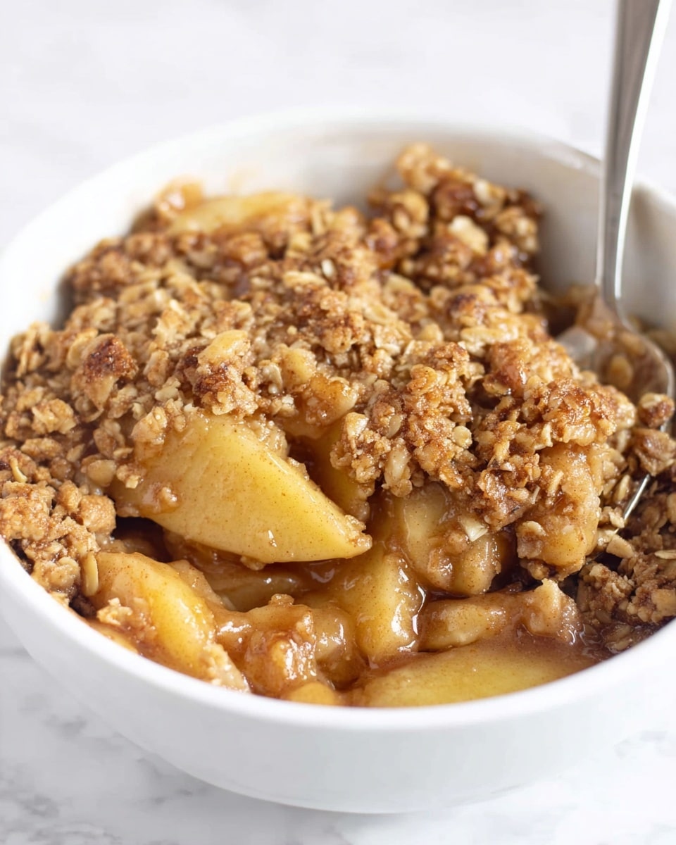 The image shows a white bowl filled with a warm apple crumble. The bottom layer has soft, cooked apple slices in a light brown syrup that looks sticky and shiny. On top of the apples is a thick layer of golden brown crumble made of oats and small nut pieces, giving a rough and crumbly texture. A silver spoon is partially dipped into the bowl, ready to scoop the dessert. The background is a white marbled surface. photo taken with an iphone --ar 4:5 --v 7