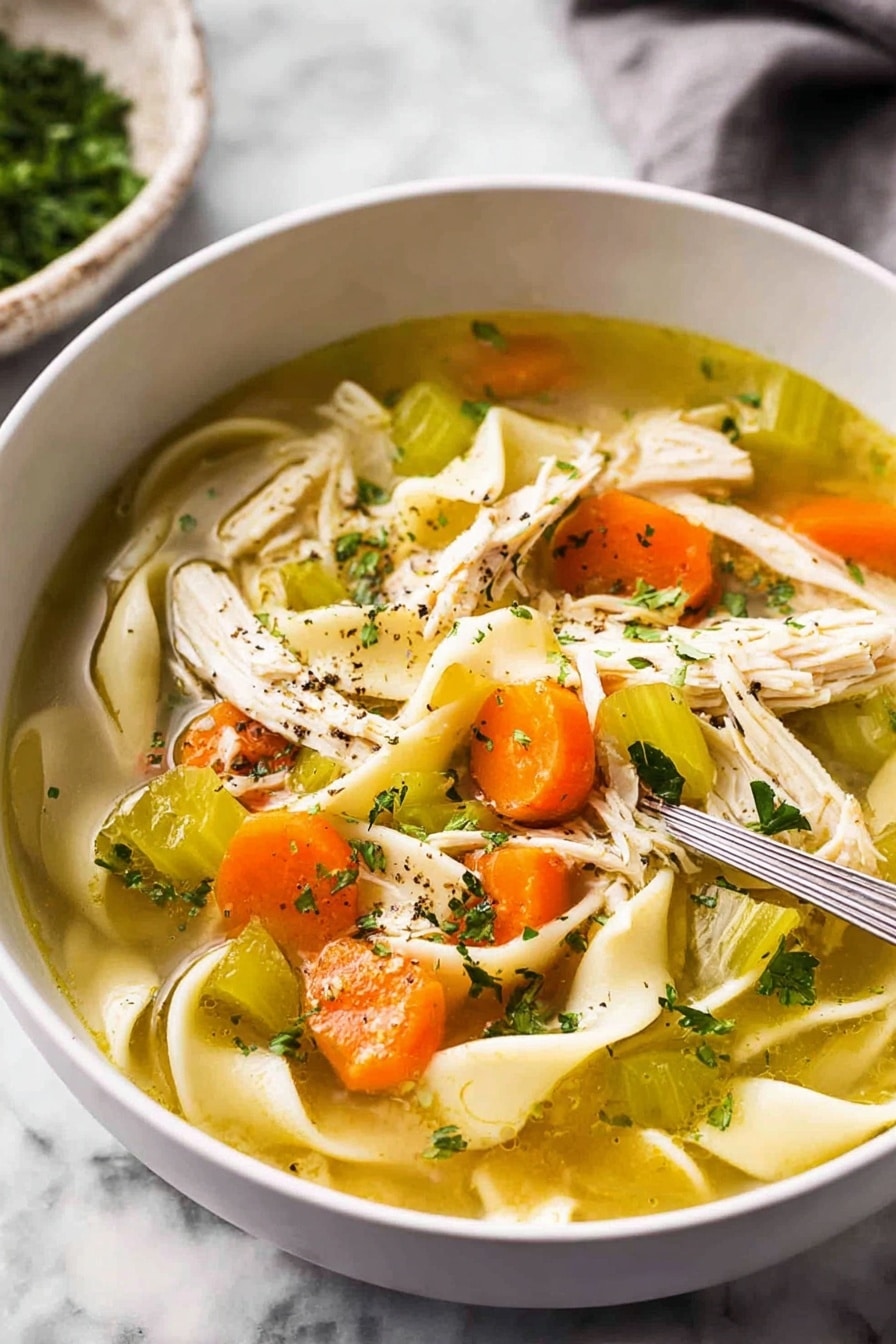 The image shows a close-up view of a white bowl filled with clear chicken soup. Inside the soup, there are wide flat noodles curled softly, bright orange carrot slices, pieces of pale green celery, and shredded white chicken meat scattered on one side. Small green parsley flakes float on the surface along with some black pepper specks. The soup is light golden and clear, with visible layers of ingredients resting naturally in the broth. The bowl sits on a white marbled surface. photo taken with an iphone --ar 2:3 --v 7 - Homemade Chicken Noodle Soup from Scratch, hearty chicken noodle soup, homemade chicken soup recipe, from scratch chicken noodle soup, comforting chicken noodle soup