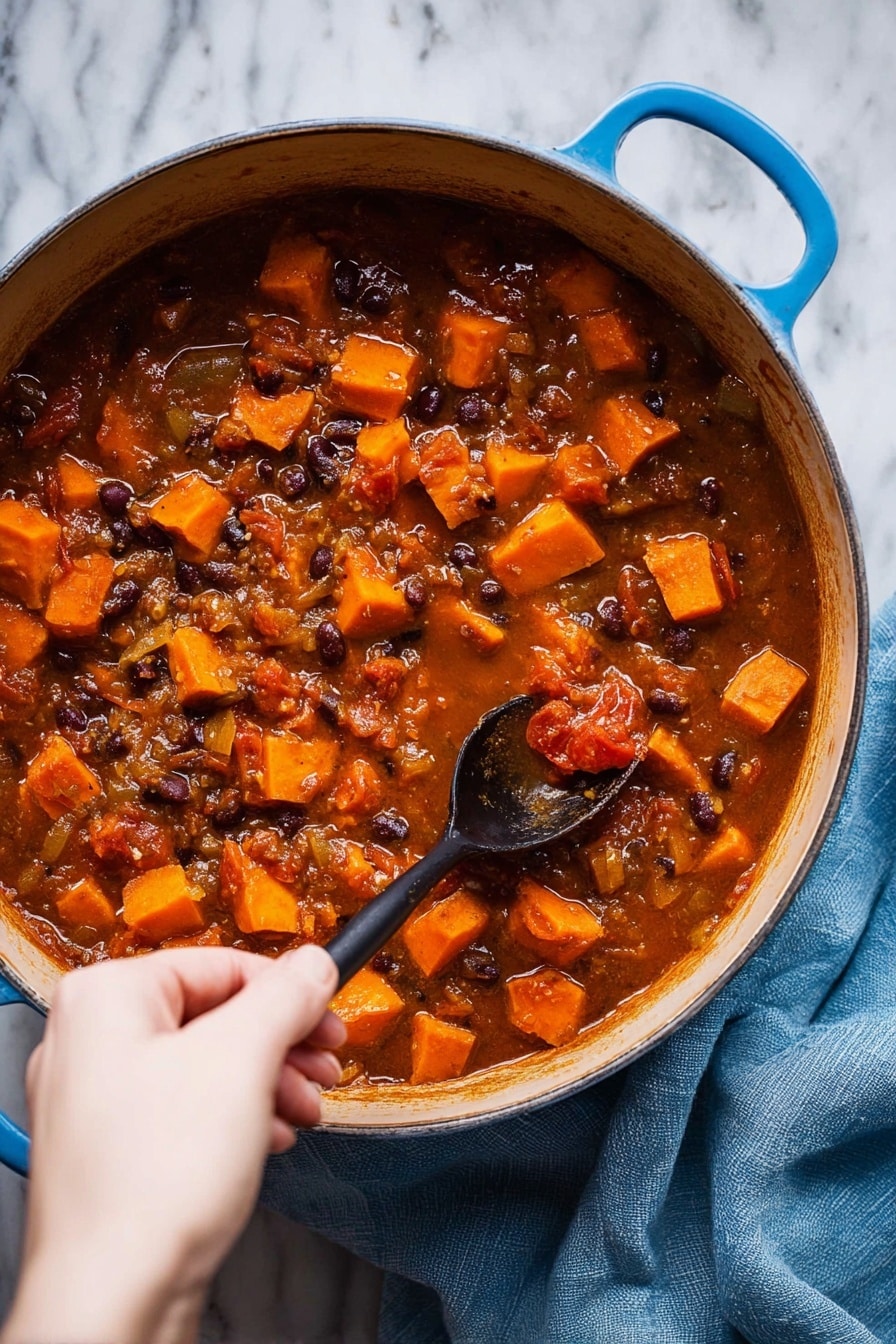 A large blue pot filled with a thick rich stew that has many small orange cubes of sweet potato, dark beans, and pieces of tomato in a brownish-red liquid. The stew has visible chunks of onion and a textured look from the cooked ingredients. A woman's hand is holding a black spoon, stirring the stew gently. The pot is placed on a white marbled surface, with a soft blue cloth nearby. Photo taken with an iphone --ar 2:3 --v 7 - Sweet Potato Black Bean Soup Ginger Lime, vegan black bean soup with sweet potatoes, nourishing vegan soup recipes, hearty plant-based soups, easy spicy veggie soup