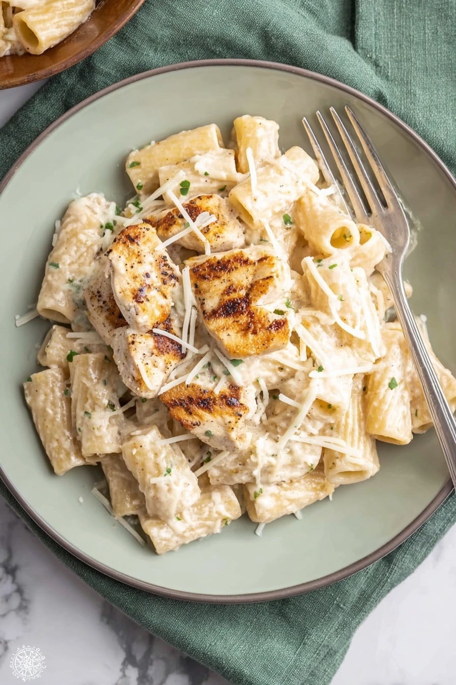 A white plate holds a serving of rigatoni pasta coated in a creamy white sauce with small green herb flecks. On top of the pasta lie four grilled chicken pieces, light brown with darker grill marks, partially covered by the sauce. Thin white shredded cheese is sprinkled over the chicken and pasta, adding texture. A silver fork rests on the right side of the plate. The plate is on a soft green cloth placed on a white marbled surface. Photo taken with an iphone --ar 2:3 --v 7 - One Pot Creamy Parmesan Chicken Pasta, creamy chicken pasta with Parmesan, easy one pot chicken pasta, cheesy chicken pasta dinner, quick creamy pasta recipe