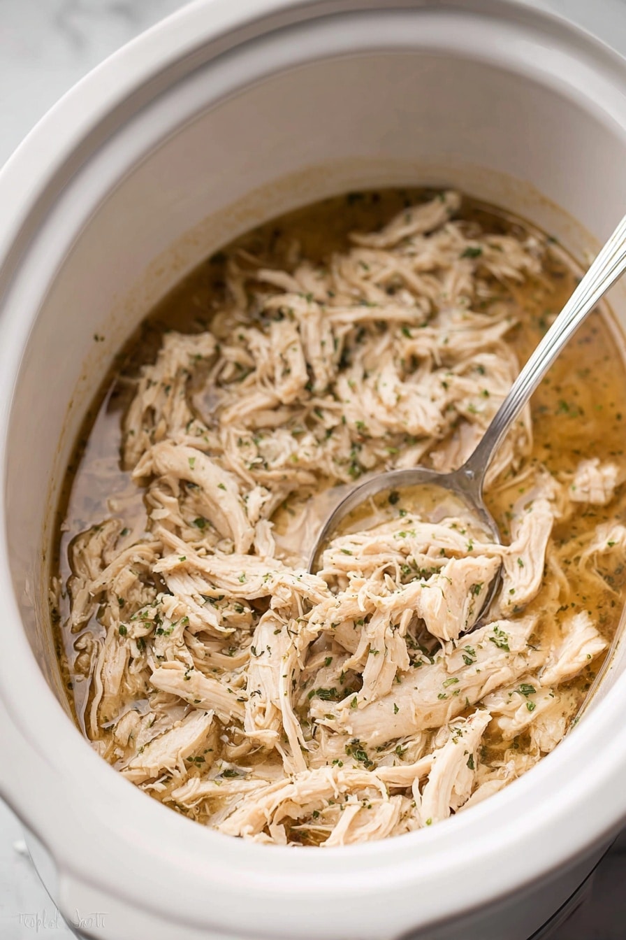Inside a white slow cooker, there is one main layer of shredded light beige cooked chicken mixed with small green herb pieces, all soaked in a thin, light brown broth. A silver spoon rests partially submerged in the mixture on the right side. The chicken strands have a soft texture, and the herbs are finely sprinkled. The slow cooker sits on a white marbled surface. photo taken with an iphone --ar 2:3 --v 7 - Juicy Crockpot Shredded Chicken, easy slow cooker chicken, tender shredded chicken recipe, effortless chicken dinner, healthy crockpot chicken