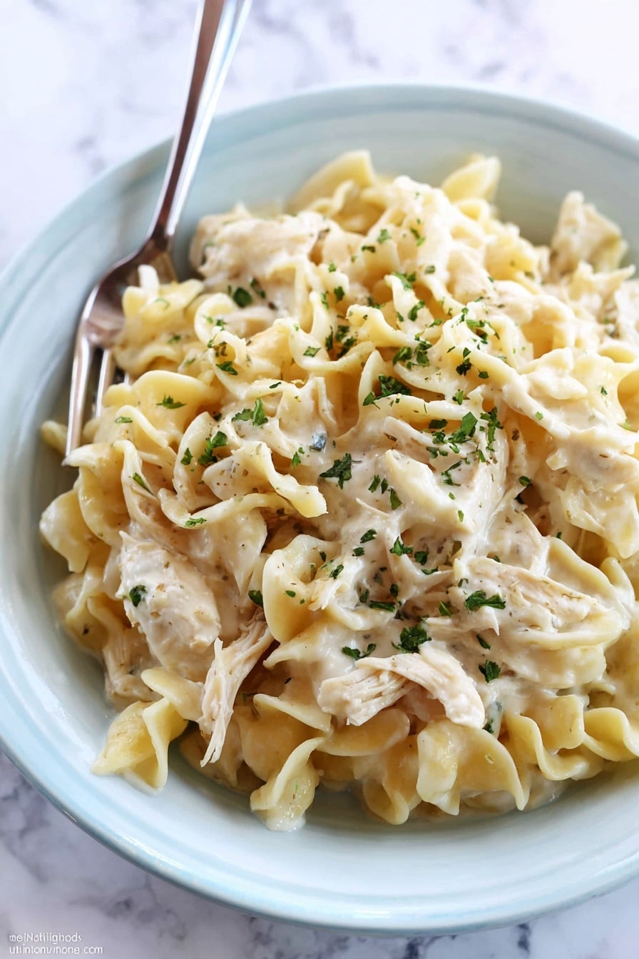A close-up of creamy chicken noodle dish served in a white bowl placed on a white marbled surface. The dish has three main layers: at the bottom is a thick white cream sauce, on top of it a layer of soft cooked egg noodles with a slightly wavy texture and pale yellow color, and scattered throughout and on top are pieces of shredded chicken mixed well with the noodles. The whole dish is sprinkled with small green herb flakes for garnish. A fork is resting inside the bowl, ready to eat. photo taken with an iphone --ar 2:3 --v 7 - Crockpot Chicken and Noodles, slow cooker chicken and noodles, homemade chicken noodle dinner, cozy crockpot chicken dish, easy chicken and noodles recipe