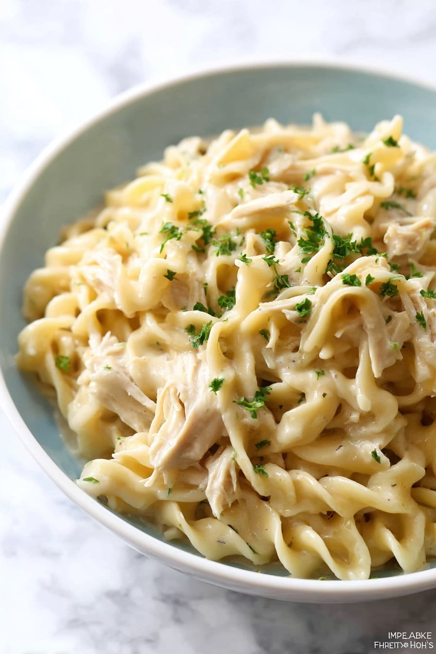 A close-up of a white bowl filled with creamy egg noodles mixed with shredded pieces of light beige chicken. The egg noodles are thick and twisted, coated in a smooth, light cream sauce. Small bits of green herbs are sprinkled on top and some are mixed within the noodles, adding small dots of green color. The bowl is set on a white marbled surface, creating a clean and bright background. Photo taken with an iphone --ar 2:3 --v 7 - Crockpot Chicken and Noodles, slow cooker chicken and noodles, homemade chicken noodle dinner, cozy crockpot chicken dish, easy chicken and noodles recipe