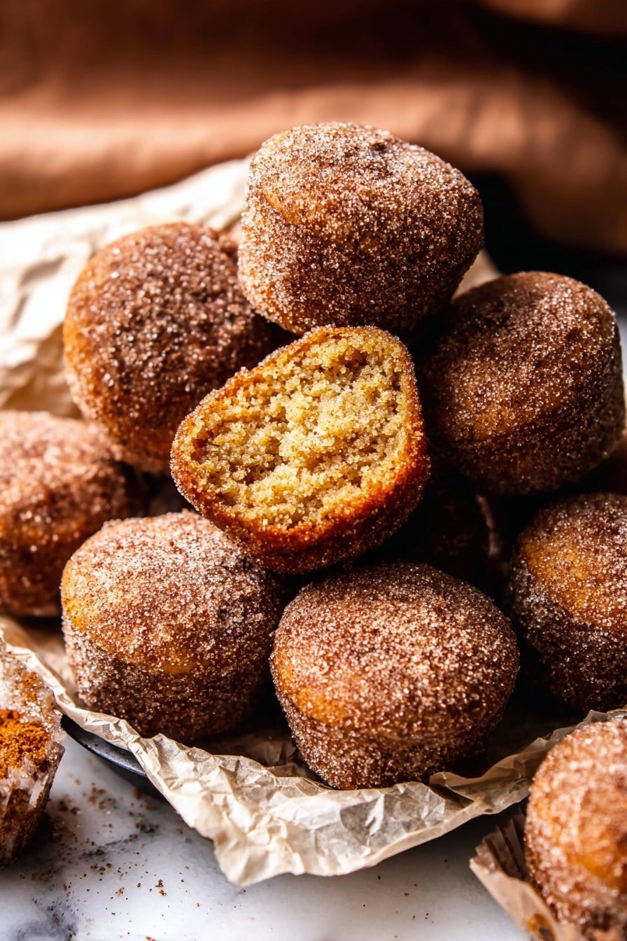The image shows a pile of small, round doughnut muffins covered in cinnamon sugar on a white marbled surface. There are about ten muffins closely stacked, with one muffin on top showing its inside layer which is soft and light brown with a slightly crumbly texture. The muffins have a rough, grainy outer layer from the sugar coating, and they are resting on crumpled white parchment paper. The scene captures a warm, cozy feeling from the brown and golden hues of the muffins, with some muffins sitting inside a black tray. photo taken with an iphone --ar 2:3 --v 7 - Baked Cinnamon Sugar Apple Cider Doughnut Holes, fall dessert recipes, cinnamon sugar donut holes, baked doughnut bites, cozy autumn treats