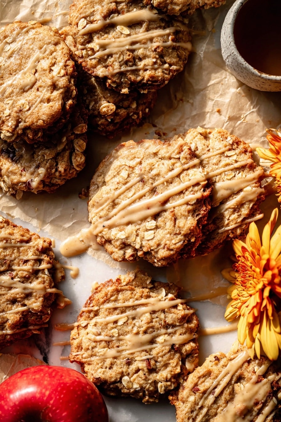 Several oatmeal cookies are spread on light brown parchment paper over a white marbled surface. The cookies are golden brown with a rough texture, showing visible oats and slightly darker edges that look crispy. Each cookie is drizzled with a thin layer of light caramel-colored glaze, creating irregular lines and patches across their tops. In the bottom left corner of the image, two whole red apples with light yellow patches are placed together, adding a fresh contrast to the warm cookie colors. The light source creates soft shadows and highlights, enhancing the cookies’ textures and the glaze's shine. photo taken with an iphone --ar 2:3 --v 7 - Iced Apple Cider Caramel Oatmeal Cookies, apple cider caramel cookies, oatmeal caramel cookies, fall cookies with caramel and oats, easy apple cider cookies