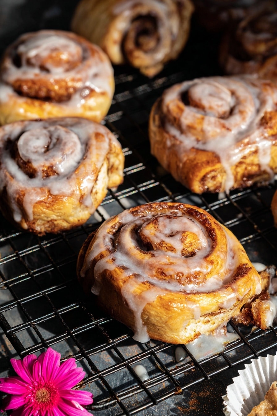 The image shows several cinnamon rolls on a black wire cooling rack with a dark base underneath. Each cinnamon roll has visible spiral layers of golden-brown dough swirled with darker brown cinnamon sugar. The top layers are coated with a shiny white glaze that drips slightly down the sides and onto the rack. The rolls have a soft, fluffy texture with a mix of light yellow and brown tones, and some darker, slightly crisped edges. Near the bottom right, there is a white paper liner with a bright pink flower placed on it. The light creates highlights on the glossy glaze and shadows between the cinnamon rolls, giving the scene depth and warmth. Photo taken with an iphone --ar 2:3 --v 7 - Shortcut Easy Apple Fritters, apple fritters with puff pastry, quick apple dessert recipes, baked apple fritters, fall-inspired apple treats