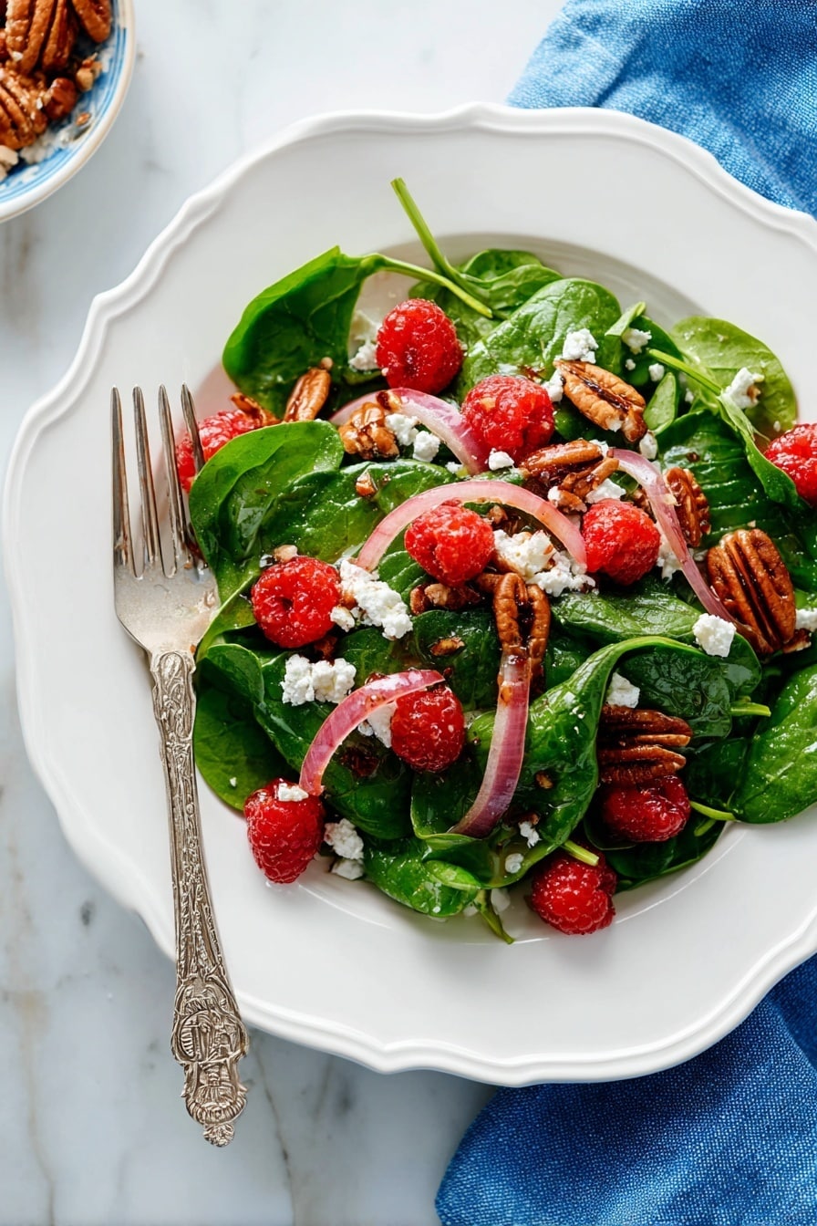 A white scalloped plate holds a fresh salad with three main layers: the base layer has bright green spinach leaves with soft texture, the middle layer has thin slices of pale pink-red onions scattered evenly, and the top layer features bright red raspberries, crumbled white cheese, and brown pecan halves arranged to create color contrast. A silver fork with an ornate handle rests on the left side of the plate, partially on the salad. The plate is placed on a white marbled surface with a blue cloth napkin nearby. Photo taken with an iphone --ar 2:3 --v 7 - Mixed Spinach Salad with Goat Cheese, Raspberries, and Toasted Pecans, spinach salad with goat cheese and raspberries, healthy berry spinach salad, easy gourmet salads, fresh fruit and nut salads