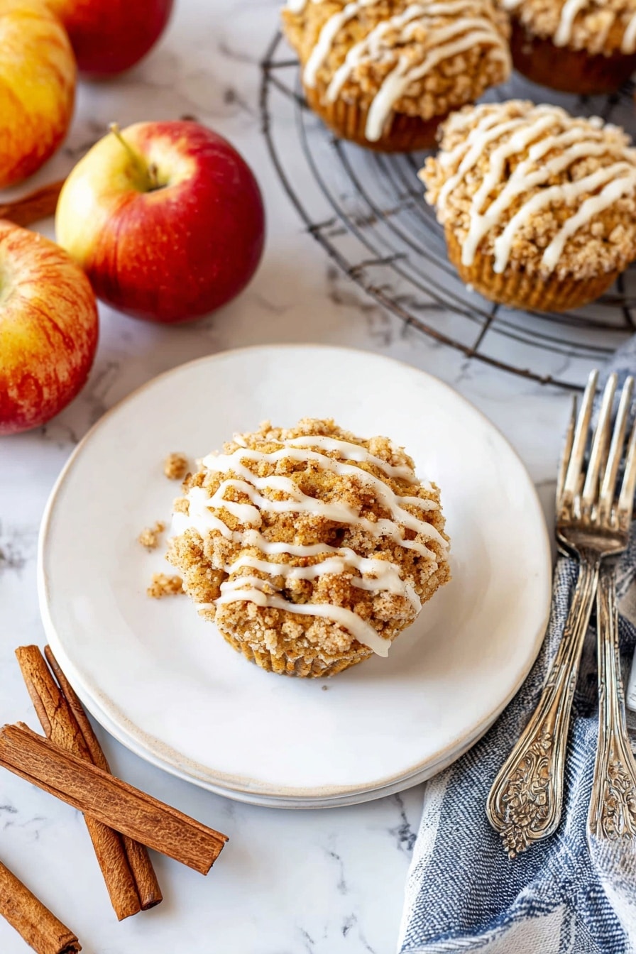 A single round muffin sits in the middle of a white plate, topped with a crumbly golden-brown streusel layer and drizzled with thick white icing in uneven stripes. The plate is on a white marbled surface surrounded by three shiny red and yellow apples, two cinnamon sticks, and a blue and white striped cloth. To the right, a wire cooling rack holds several more muffins with the same crumb topping and white icing drizzle. Two ornate silver forks rest beside the plate. The scene is bright and cozy, with warm autumn colors and textures. photo taken with an iphone --ar 2:3 --v 7 - Cinnamon Apple Muffins with Crumble Topping and Glaze, fall breakfast muffins, spiced apple muffins, easy apple muffins recipe, cozy morning muffins