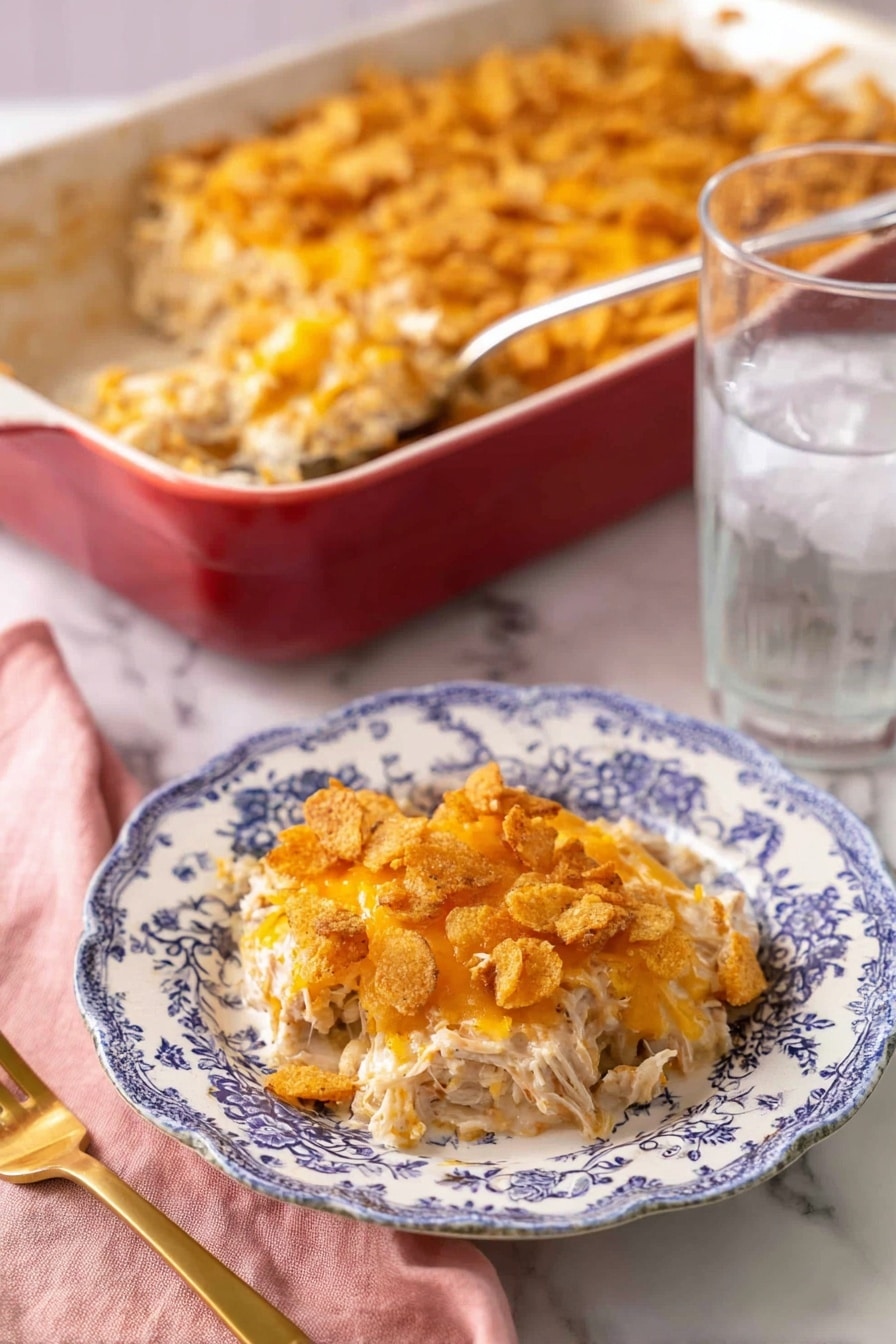 A portion of a creamy casserole is served on a white plate with blue floral patterns, showing three main layers: the bottom layer is shredded white meat mixed with a light creamy sauce, the middle layer has melted orange cheese blended with the meat, and the top layer consists of crushed crispy crackers with more melted cheese giving a golden-brown, crunchy texture. Behind the plate, there is a large red baking dish filled with the same casserole, with a silver serving spoon inside. To the right of the plate is a clear glass of ice water, and to the left is a gold fork resting on a pink cloth napkin, all placed on a white marbled surface. photo taken with an iphone --ar 2:3 --v 7 - Creamy Chicken Casserole, Chicken casserole recipe, comfort food dinner, easy chicken casserole, cheesy chicken casserole
