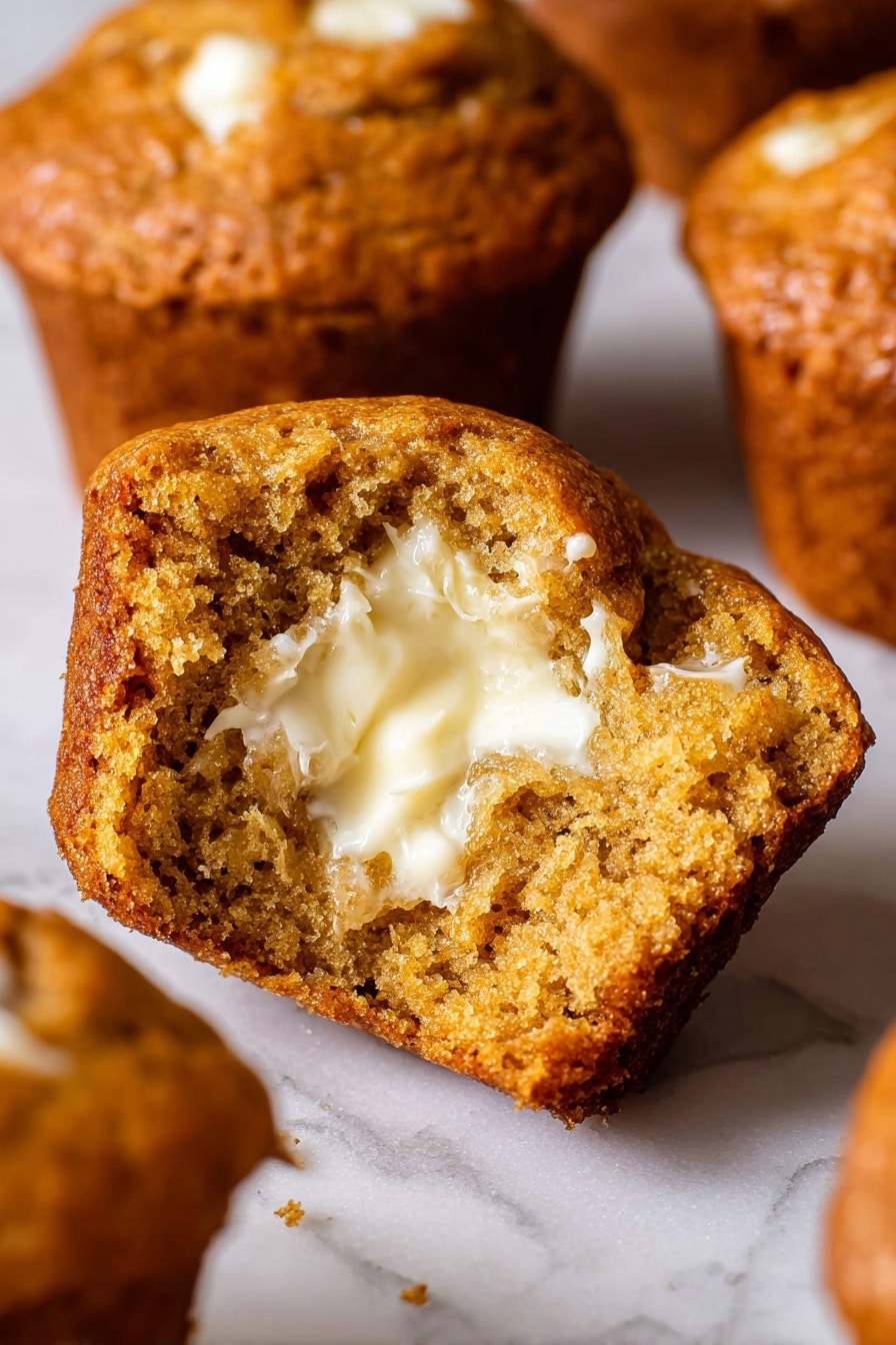 The image shows a close-up of a muffin broken in half, revealing a soft, moist texture inside with a spread of creamy, melted butter on one side. The muffin’s outer crust is rich golden brown and slightly crisp, while the inside is a dense, honey-brown color with small air pockets. Surrounding the main muffin piece, there are other muffins blurred slightly in the background, all with a similar warm golden outer color. The surface beneath the muffins is a white marbled texture. photo taken with an iphone --ar 2:3 --v 7 - Fluffy Pumpkin Muffins with Warm Spices, pumpkin muffin recipe, autumn pumpkin muffins, cozy fall muffins, soft pumpkin spice muffins